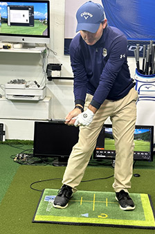 A person in golf attire practices their swing indoors on a golf mat, holding a club and wearing a golf glove. Several screens and golf equipment are visible in the background.