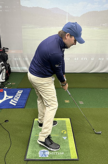 A golfer in a blue jacket and khaki pants prepares to take a shot indoors, standing on a sensor mat with a golf club in hand. A golf simulator screen with a course image is displayed in the background.