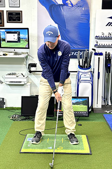 A golfer in a blue shirt and khaki pants stands indoors on a practice mat, preparing to swing a golf club, with golf equipment and monitors visible in the background.