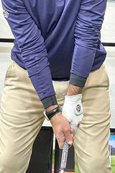 A person wearing a blue long-sleeve shirt and beige pants is gripping a golf club, preparing to swing. Only the torso and arms are visible.
