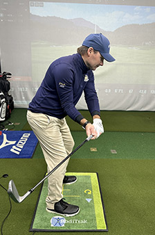 A man wearing a blue cap, blue jacket, and beige pants prepares to swing a golf club indoors, standing on a mat with golf equipment and a virtual golf screen in the background.