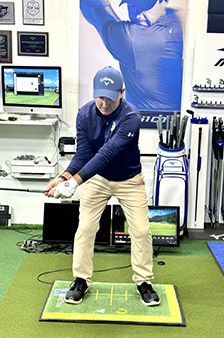 A man wearing a blue cap and shirt, beige pants, and black shoes stands in a golf stance on an indoor practice mat, holding a golf club. Golf equipment and monitors are visible in the background.