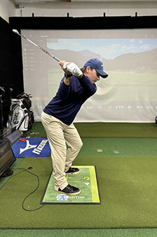 A person wearing a blue cap and sweater with beige pants prepares to swing a golf club indoors, standing on an artificial green mat with a screen displaying a virtual golf course in the background.