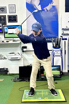 A man in a golf studio stands in a backswing position on a golf mat, holding a golf club. He wears a blue cap, blue long-sleeve shirt, and beige pants. Golf equipment and a large golf poster are in the background.
