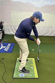 A man wearing a blue cap, navy shirt, and beige pants stands in a golfing stance indoors on a green mat, holding a golf club, with golf equipment and a simulator screen in the background.