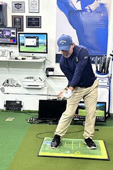 A man in golf attire practices his golf swing indoors on a green mat, surrounded by golf equipment, computers, and monitors displaying golf-related data in a training facility.