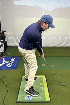 A man wearing a blue cap, blue jacket, and khaki pants holds a golf club in a ready stance indoors on a green mat, with golf equipment and a projected course screen in the background.