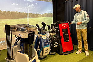 tile-club-fitting2 A man in a golf cap and sweater stands indoors beside several golf bags and a red Callaway club fitting cart, with a golf course scene projected on a screen behind him.