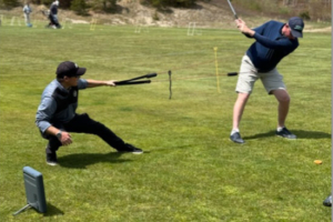 tile-full-development A golf instructor crouches and uses training aids to guide a golfer’s swing on a grassy practice range, with golf equipment and targets visible in the background.
