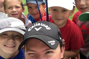 tile-junior-clinics A group of smiling children and one adult in a Callaway cap take a close-up selfie outdoors, possibly at a golf event, with green grass in the background.