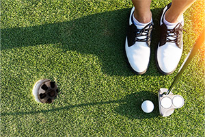 tile-lesson-gift-cards A person wearing black and white golf shoes stands on green grass next to a golf hole, preparing to putt a golf ball with a putter.