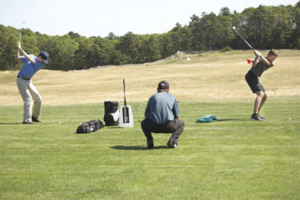 tile-private-lessons Three people are on a golf course; two are preparing to swing golf clubs, while the third crouches between them, observing. Golf bags and equipment are nearby on the grass. Trees and hills are in the background.