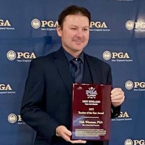 A man in a dark suit holds a red plaque while standing in front of a blue backdrop with multiple PGA logos. The plaque reads Teacher of the Year Award. He is smiling for the photo.